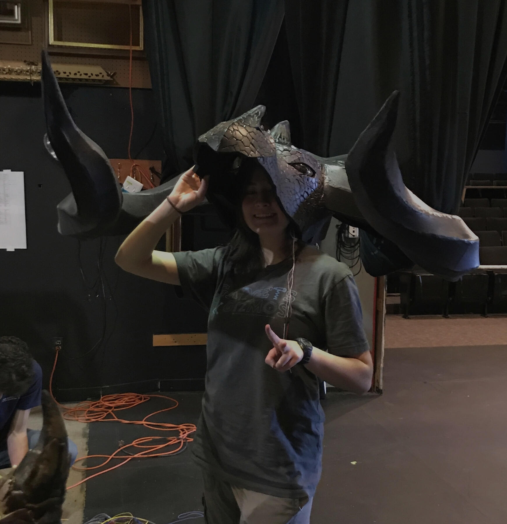 Picture of natasha backstage, dwarfed by the massive black dragon head which is on their head like a hat. They are smiling and holding up a peace sign